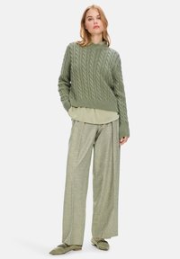 Grüner, aus Kabelstrick gefertigter, cropped Pullover über einer hellgrünen Bluse, kombiniert mit weit geschnittenen, hellgrünen gemusterten Hosen und passenden Schuhen.