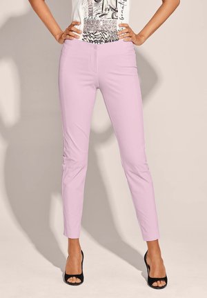 Slim Fit Lavendelhose kombiniert mit schwarzen offenen Absätzen, getragen mit einem weißen grafischen, in die Hose gesteckten Oberteil, stehend mit Händen in der Taille.