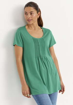 Kvinde, der læner sig op ad en hvid væg iført en grøn top med korte ærmer med lodrette folder og lyseblå jeans, ser mod venstre.