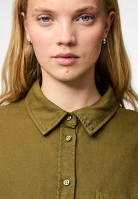 Olivgrön button-up skjorta med krage, med texturerad tyg och guldknappar, visad mot en neutral bakgrund.