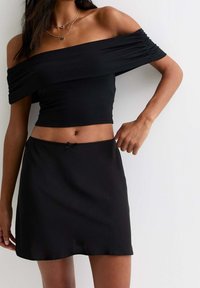 Mujer vistiendo un top corto negro de hombros al aire y una falda negra a juego, mostrando el abdomen con collares en capas, contra un fondo blanco liso.