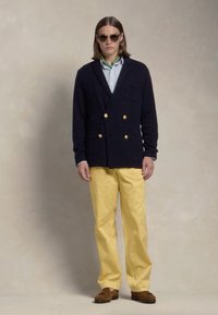 Cardigan dublu încheiat, de culoare bleumarin, cu butoane aurii, cămașă în dungi dedesubt, asortat cu pantaloni galbeni și mocasini maro. Ochelari de soare purtați.
