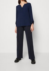 Femme portant un chemisier bleu marine à manches longues avec des boutons et un pantalon large à fines rayures, assorti à des chaussures noires épaisses.