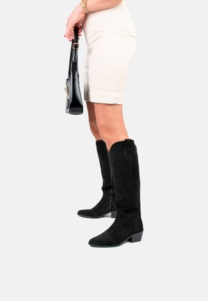 Schwarze Wildleder-Stiefel bis zum Knie mit einer spitzen Zehenform und einem Blockabsatz; glatte Textur und minimalistisches Design. Kombiniert mit einem hellbeigen Rock.