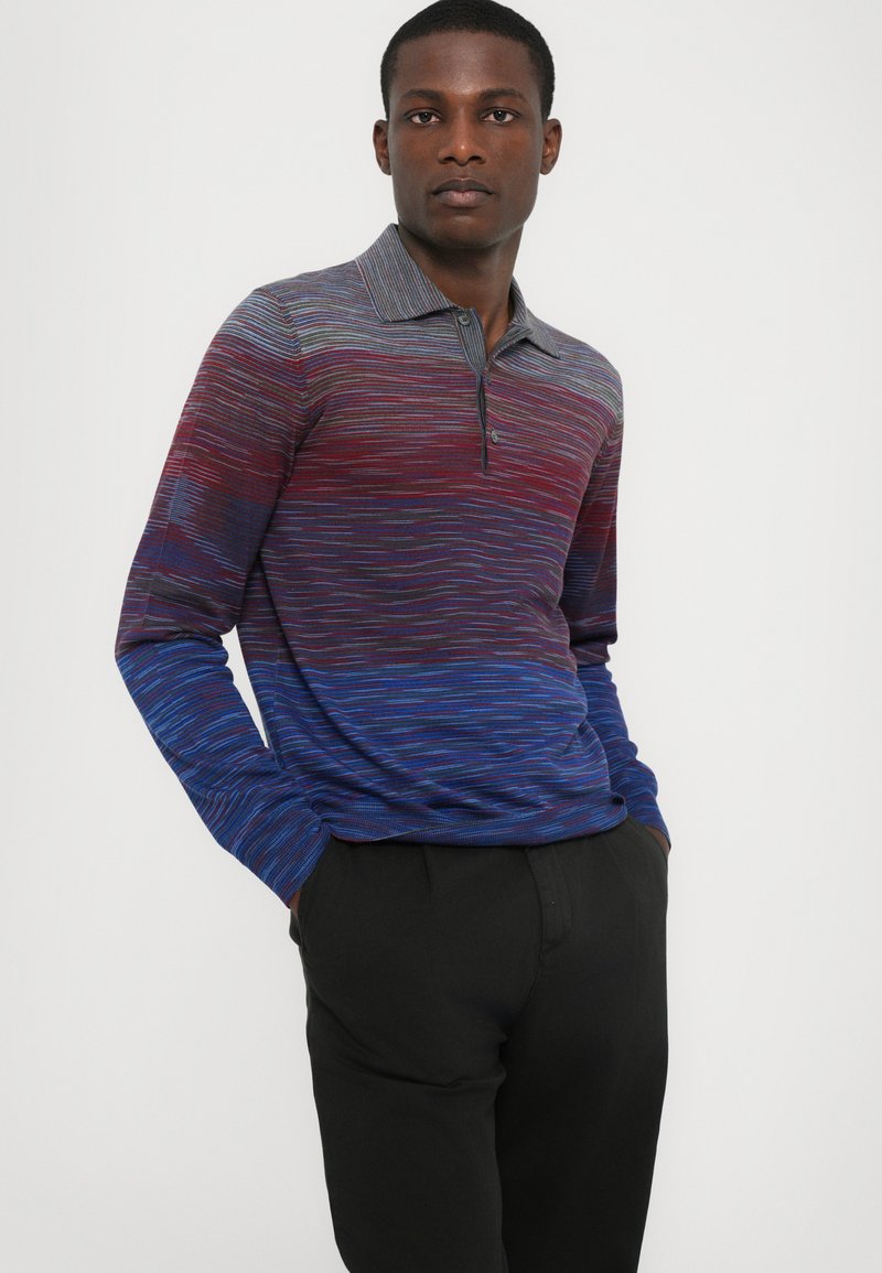 Polo de manga larga en rayas azul, roja y púrpura, con cuello plano y tres botones. Textura de punto suave. Pantalones negros.