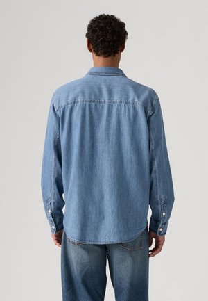 Homme portant une chemise en denim bleu clair et un jean, debout de dos devant un fond uni clair.