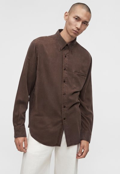 Νέος άνδρας με κοντά μαλλιά, που φοράει σκούρο καφέ πουκάμισο με κουμπιά και λευκό παντελόνι, στέκεται μπροστά σε απλό φωτεινό φόντο.