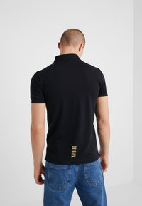 Svart polo skjorta med en liten gul rand baktill, kortärmad och med klassisk krage. Bärs med blå denimjeans.