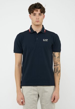 Joven con cabello corto y rizado, vistiendo una camiseta polo azul marino EA7 y pantalones beige, mostrando tatuajes en su antebrazo derecho.