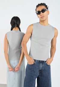 Grå ermeløs topp med ribbestruktur, paret med blå denimjeans. To modeller står rygg mot rygg, den ene med solbriller og den andre med flette.
