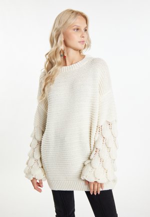 Oversized cream sweater with textured, scalloped knit sleeves. Ribbed neckline and hem, paired with fitted black pants.