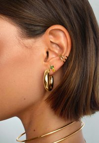 Pendiente de aro dorado y pequeño pendiente de piña verde y amarilla en una oreja perforada. Textura suave, acabado brillante y diseño moderno.