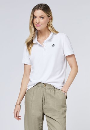 Weißes Poloshirt mit Kragen, einer dreiknöpfigen Knopfleiste und einem kleinen schwarzen Logo auf der linken Brust; kombiniert mit hellen Khakihosen.