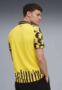Cămașă polo galbenă cu mâneci scurte cu model maroniu și tiv inferior. Prezintă un guler negru și branding discret în spate.