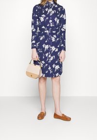 Mörkblå blommig klänning med långa ärmar och knappar, bältad i midjan. Håller en liten handväska i tan och rosa, kombinerad med bruna loafers.