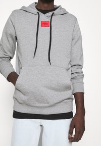 Sudadera gris con un bolsillo tipo canguro, cordones negros y un notable parche de logo rojo "HUGO" en el pecho. Tejido suave y texturizado.
