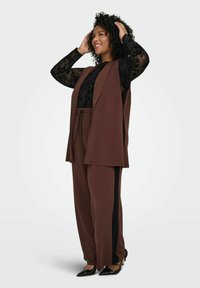 Nasmejana žena prilagođava kovrčavu kosu, nosi smeđe široke hlače, odgovarajući bezrukavni sako, crnu čipkastu bluzu s dugim rukavima i crne cipele s dugim vrhom.