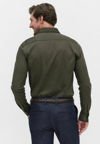 Chemise à manches longues vert foncé avec une texture lisse, des poignets à boutons et un col, portée avec un pantalon bleu marine et une ceinture tressée marron.