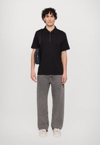 Svart kortermet polo med høy krage og glidelås, kombinert med lysegrå baggy jeans og uformelle beige joggesko. Standard skulderveske.