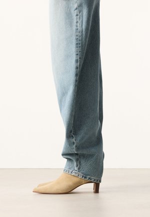 Lichtblauwe spijkerbroek gedragen met beige suède puntige kitten heel schoenen, zijwaarts afgebeeld tegen een effen achtergrond.