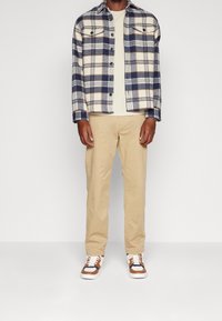 Camicia di flanella a quadri in blu e crema con tasche sul petto con bottoni, maglietta beige chiaro a girocollo, pantaloni khaki e sneakers multicolori.