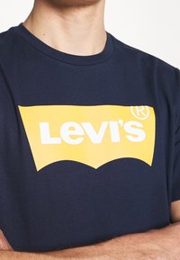 Námorně modré bavlněné tričko s velkým žlutým logem Levi's a bílým textem, s klasickým kulatým výstřihem a krátkými rukávy.
