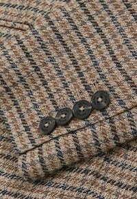 Hnedá a béžová látka s vzorom houndstooth a textúrovanou väzbou. Štyri čierne gombíky s odtlačkom značky na manžete.