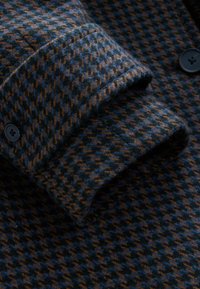 Wool shirt featuring a houndstooth pattern in blue and brown, with a buttoned cuff and a textured finish.
