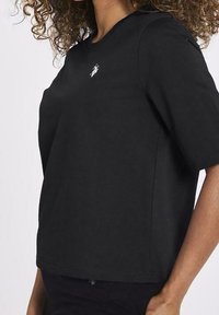 Schwarzes Rundhals-T-Shirt mit kurzen Ärmeln, aus weichem Stoff gefertigt. Enthält ein kleines gesticktes Logo auf der oberen linken Brust.