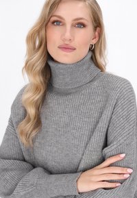 Grijze geribbelde coltrui met lange mouwen, voorzien van een gestructureerde stof en een brede kraag. Het model heeft zachte golven in haar haar.