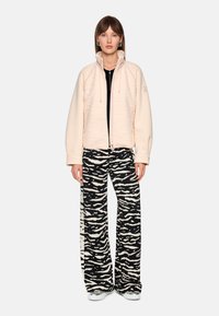 Light pink zip-up jacket with textured design, paired with black and white zebra-patterned wide-leg pants. Model standing on white background.