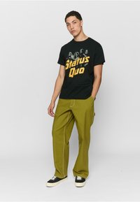 Sort t-shirt med "Status Quo" grafik; grønne bukser med synlige sømme; sorte sneakers med hvide såler; afslappet pasform.
