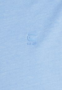 Gaiši zila kokvilnas T-kreklus ar ribotu tekstūru un reljefu logotipu, kurā centrā ir "G" un "RAW". Vienkāršs, vienkrāsains dizains.