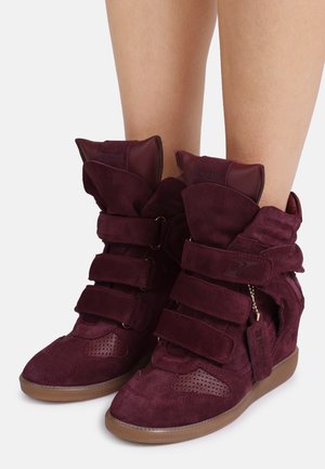 Sneakers con zeppa in camoscio color prugna con tre ampie strisce in velcro, suole di gomma marroni e un'etichetta appesa alla scarpa destra, indossate su gambe nude.