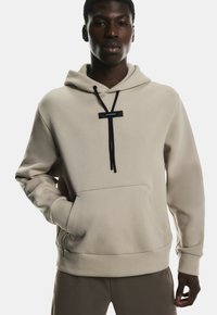 Sweat à capuche beige en tissu doux avec poche kangourou, cordons de serrage noirs et patch logo au niveau du col. Présente des poignets côtelés.