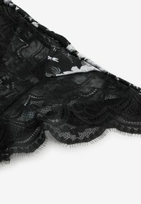 Schwarzer Spitzenstoff mit floralen Mustern und gewellten Kanten, teilweise gefaltet, um das schwarz-weiße florale Innenfutter mit Branding-Text zu zeigen.