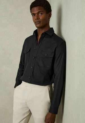 Zwarte lange mouwen shirt met geknoopte kraag, twee borstzakken, geweven stof, gecombineerd met lichtbeige broek.