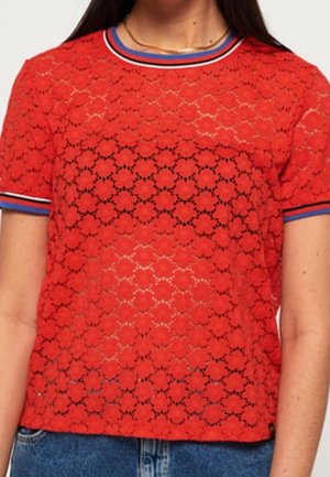 Czerwony top z krótkim rękawem z koronki w kwiatowy wzór, z niebieskim, białym i czerwonym paskowanym okrągłym dekoltem oraz mankietami na rękawach, noszony z niebieskimi dżinsami.