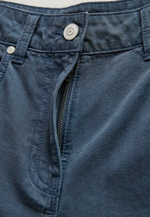 Prim-plan cu blugi albastru închis, cu fermoar parțial deschis și nasture metalic inscripționat "NEXT COLLECTION."