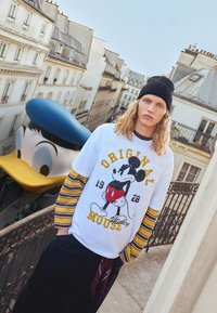 Bijela majica dugih rukava s rukavima u crno-žutim prugama koja sadrži grafiku Mickey Mousa; nosi se uz tamne hlače i crnu beanie.