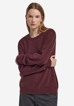 Femme aux cheveux blonds lisses portant des boucles d'oreilles en or et un pull bordeaux ample à manches longues, debout les bras croisés.