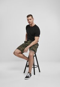 Camiseta negra de manga corta combinada con pantalones cortos de estampado de camuflaje, sentado en un taburete negro, usando zapatillas negras y blancas.