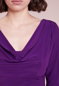 Lila klänning med en draperad halsringning och trekvartsärmar. Tyget verkar slätt med subtil textur och en lätt lyster.