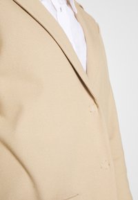 Blazer beige con un tessuto testurizzato, design a un petto, rever a punta, due bottoni frontali e una camicia bianca liscia sotto.
