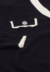 Cardigan blu navy con texture e dettagli crema, caratterizzato da un colletto scallop e una spilla gioiello sulla tasca del petto. Polsini a coste.