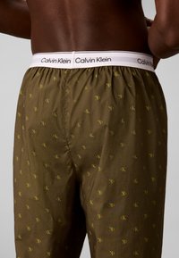 Άνδρας που φοράει σορτς μπόξερ Calvin Klein σε ελαιώδες πράσινο με μικρά κίτρινα λογότυπα "cK" και λευκή ζώνη μέσης με μαύρο κείμενο Calvin Klein.