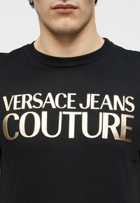 Κοντινό πλάνο ενός ατόμου που φοράει μαύρο μπλουζάκι με μεγάλα χρυσά μεταλλικά γράμματα στο στήθος με την επιγραφή "VERSACE JEANS COUTURE".