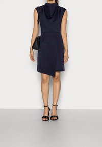 Kvinna i en ärmlös marinblå klänning med asymmetrisk nederkant, svarta klackar med ankelsnörning och en svart handväska över axeln.