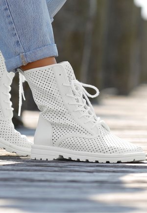 Bottines blanches perforées avec un design à lacets, surface texturée et semelle en caoutchouc épaisse, associées à un jean bleu clair retroussé.