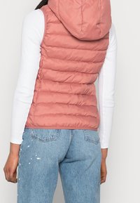 Rosa pufferväst med huva, med kviltade sektioner och en slät textur. Kombinerad med en långärmad vit topp och ljusblå jeans.